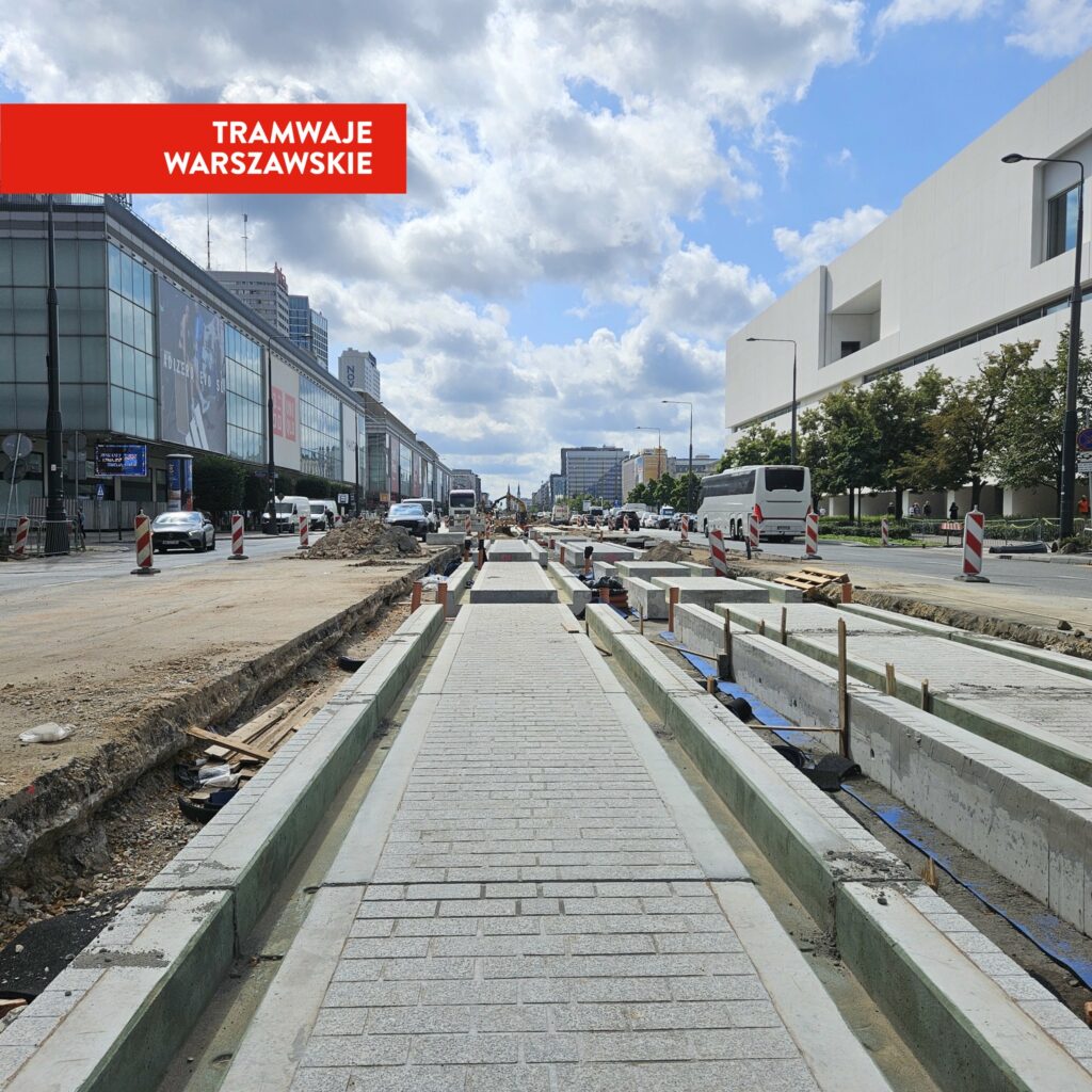 Budowa torowiska tramwajowego na ulicy, z betonowymi elementami w trakcie układania, w tle nowoczesne budynki.