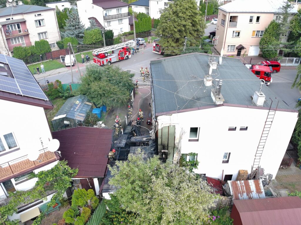 Strażacy gaszą pożar w budynku mieszkalnym, obok stoją wozy strażackie oraz zebrani mieszkańcy.