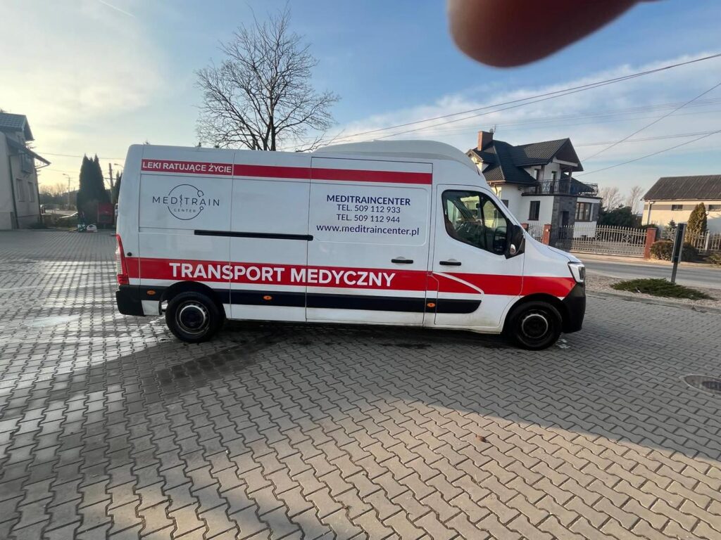 Furgonetka z napisem "TRANSPORT MEDYCZNY" zaparkowana na kostce brukowej w otoczeniu domów.