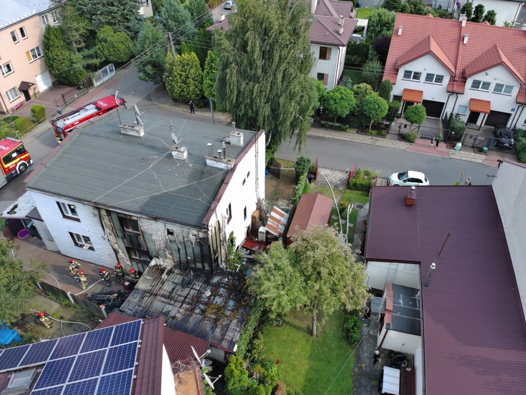 Strażacy pracują przy zniszczonej elewacji budynku mieszkalnego, obok stoją wozy strażackie na osiedlowej ulicy.