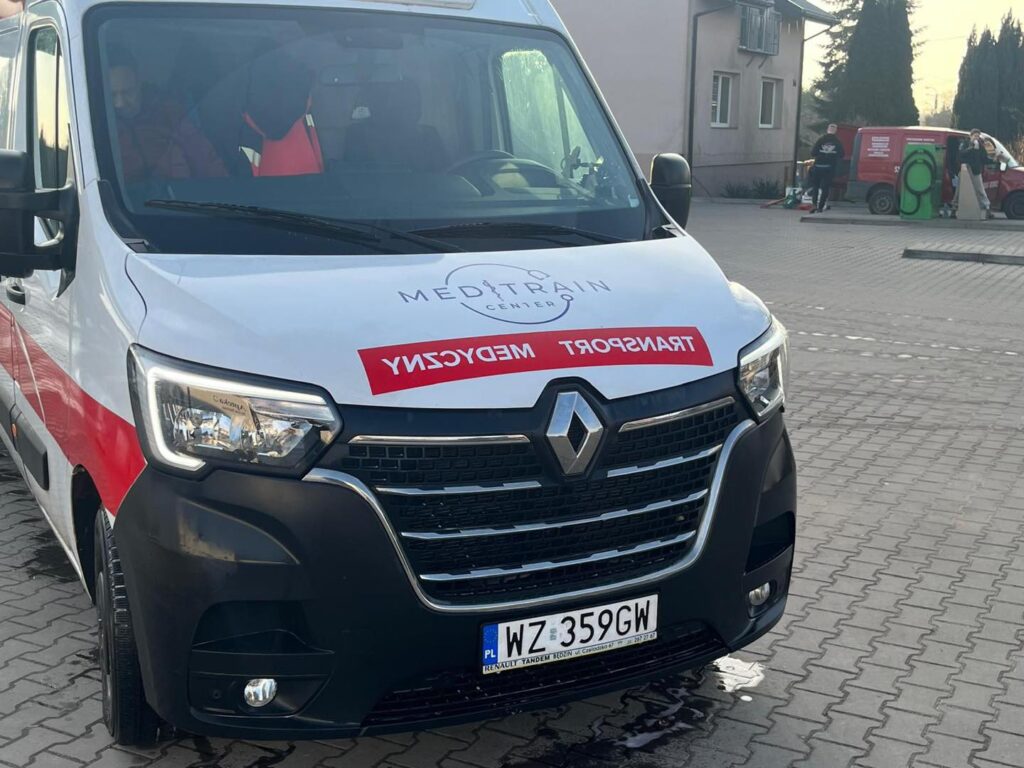 Renault w kolorach białym i czerwonym z napisem "TRANSPORT MEDYCZNY" na przodzie, zaparkowany na placu.
