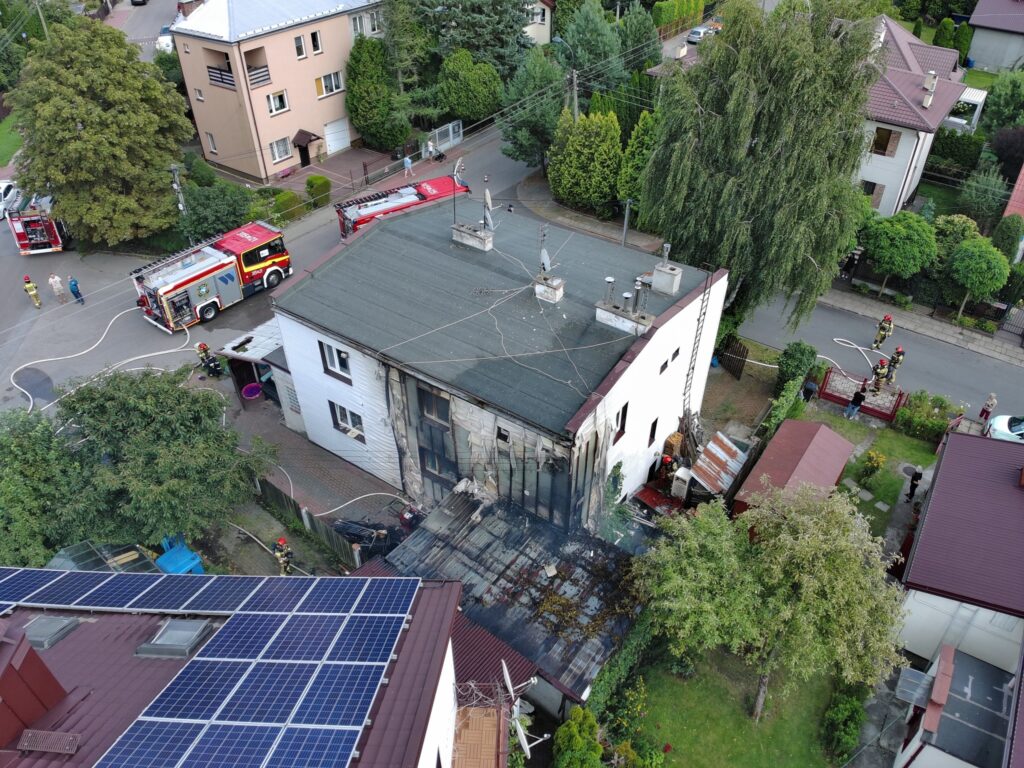 Strażacy gaszą pożar budynku mieszkalnego, obok stoją wozy strażackie, a w tle widać sąsiednie domy i zieleń.