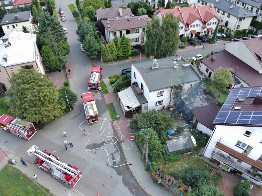 Strażacy gaszą pożar w uszkodzonym budynku mieszkalnym, otoczeni przez pojazdy strażackie na osiedlowej ulicy.