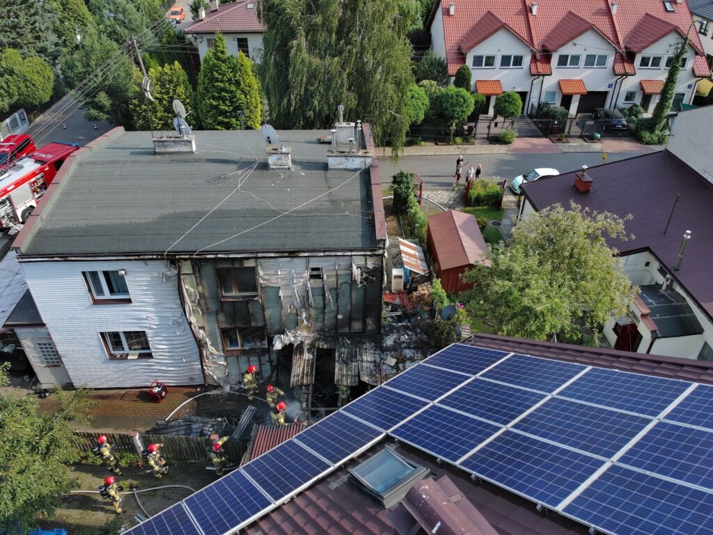 Strażacy gaszą pożar w uszkodzonym budynku mieszkalnym, otoczonym przez sąsiednie domy i roślinność.