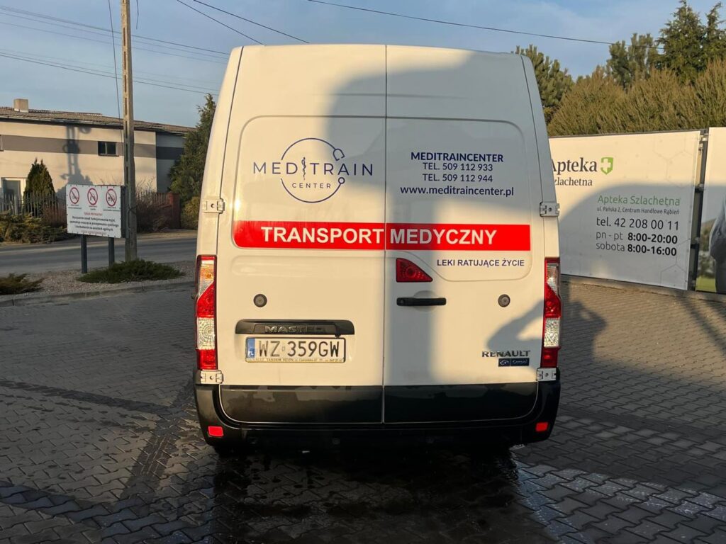 Biały samochód dostawczy z napisem „TRANSPORT MEDYCZNY” stoi na parkingu obok apteki.