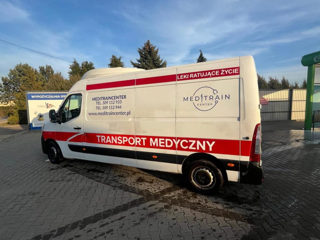 Samochód dostawczy z napisem "TRANSPORT MEDYCZNY" zaparkowany na placu, w tle widoczne drzewa i budynki.
