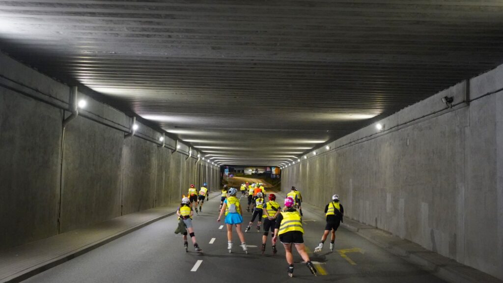 Grupa osób na rolkach porusza się przez oświetlony tunel, nosząc kamizelki odblaskowe.