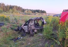 Zniszczony samochód leży w trawie obok drogi, otoczony dziką roślinnością i ogrodzeniem.