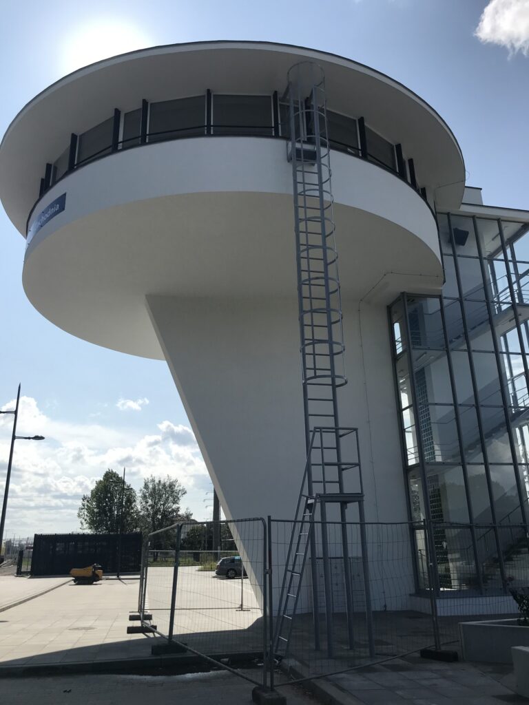 Nowoczesny budynek z okrągłym dachem i przeszkloną fasadą, obok metalowa drabina i ogrodzenie budowlane.