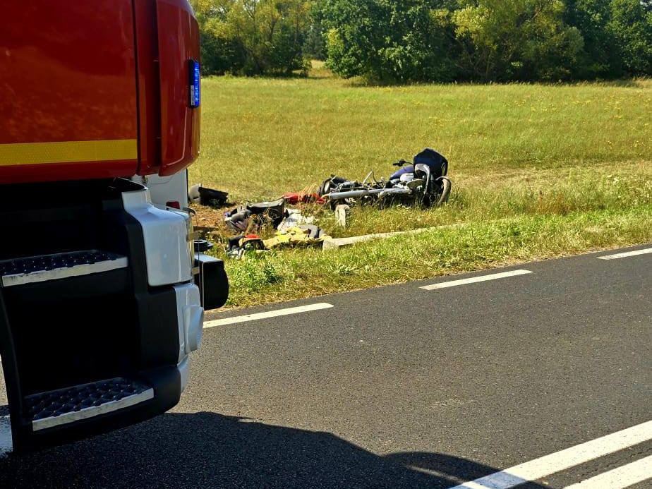 Na poboczu drogi leży przewrócony rower oraz porozrzucane przedmioty, obok stoi wóz strażacki.