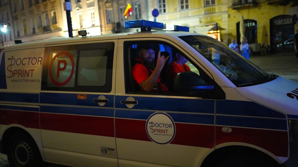 Mężczyzna w czerwonej koszulce siedzi w samochodzie z logo "DOCTOR SPRINT", machając ręką. Nocne miasto w tle.