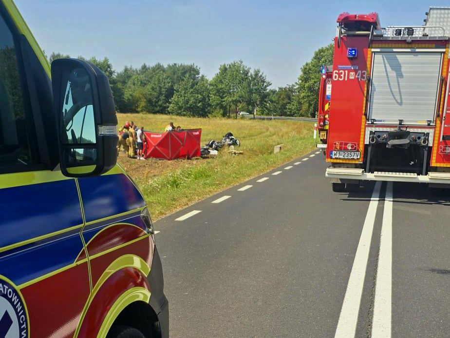 Zespół ratowników medycznych pracuje na poboczu drogi, obok czerwoną namiotu, w tle widoczny wóz strażacki.