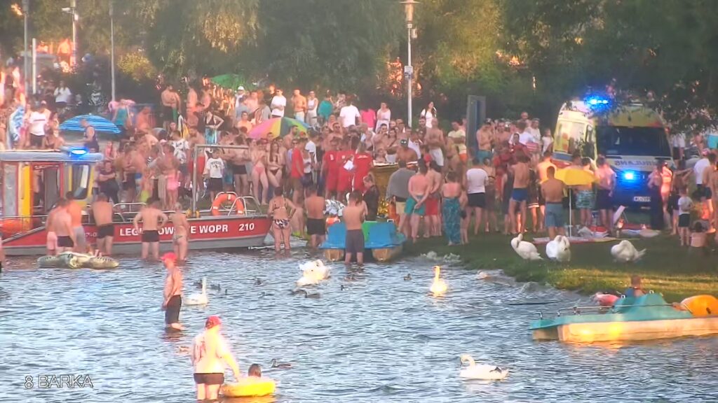 Tłum ludzi w strojach kąpielowych bawi się nad wodą, obok łodzi ratunkowej i policyjnego samochodu.