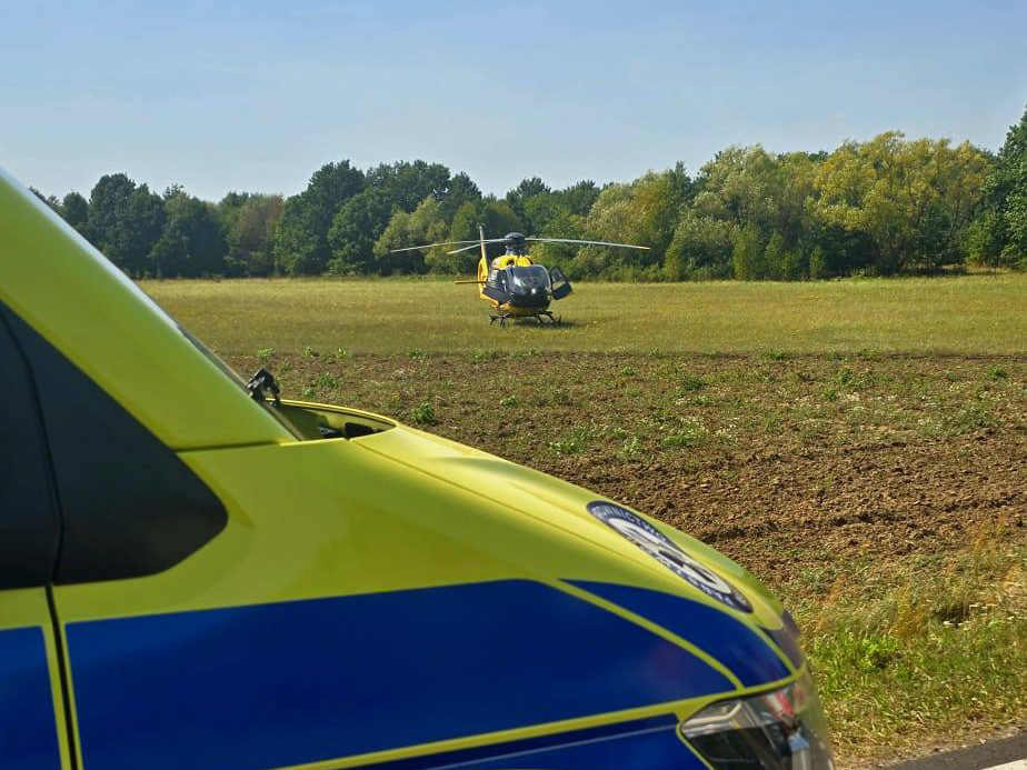 Śmigłowiec ratunkowy ląduje na polu, w tle widać zieleń drzew i nowoczesny pojazd służb ratunkowych.