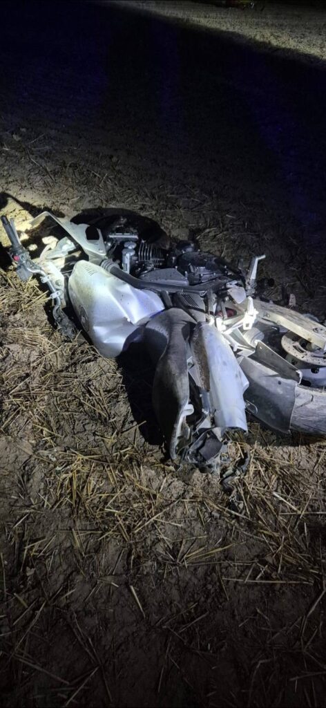 Zniszczony motocykl leży na ziemi w polu, oświetlony niebieskim światłem.