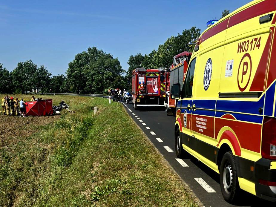 Na drodze stoją pojazdy ratunkowe, a strażacy pomagają poszkodowanym w wypadku. W tle widać zieleń drzew.