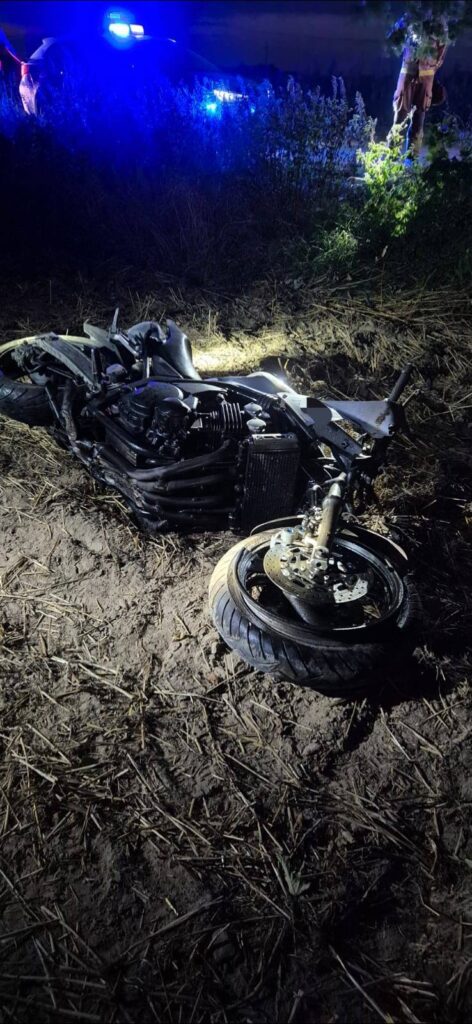 Motocykl leży na boku w zaroślach, w tle widoczne niebieskie światła policyjne.