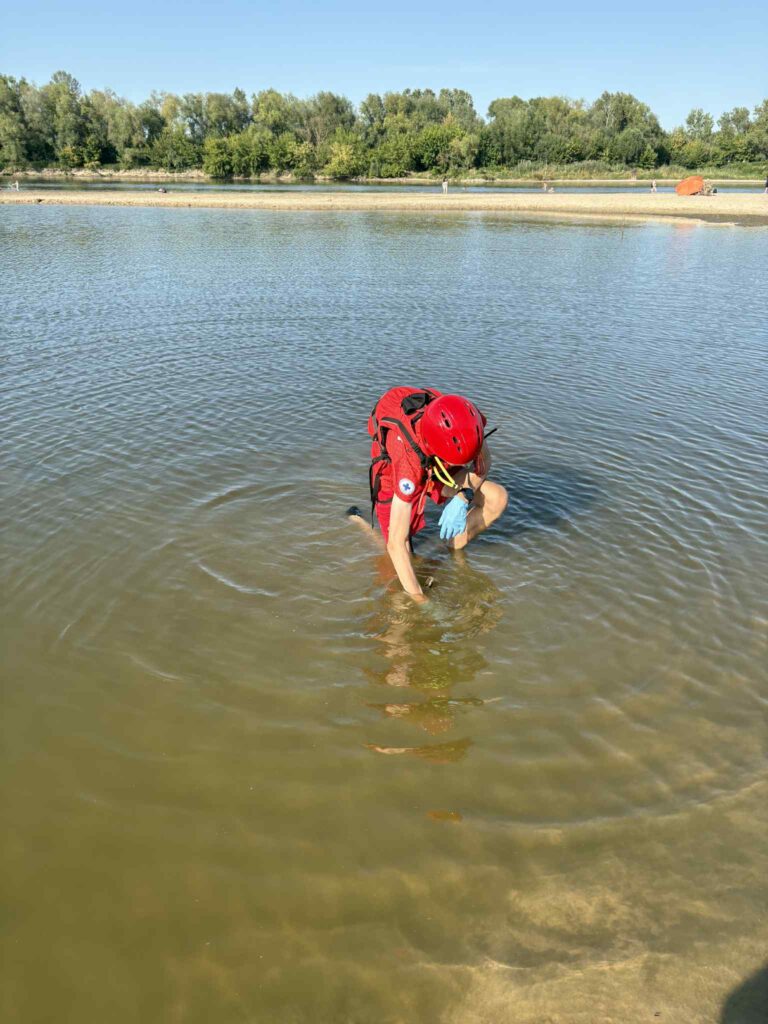 Ratownik w czerwonym uniformie sprawdzający dno w płytkiej wodzie, w tle zieleń drzew i plaża.
