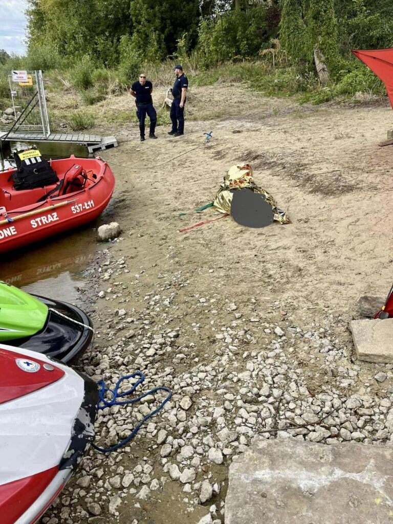 Dwaj mężczyźni w mundurach straży pożarnej stoją obok plaży, obok leżącego na ziemi obiektu przykrytego materiałem.