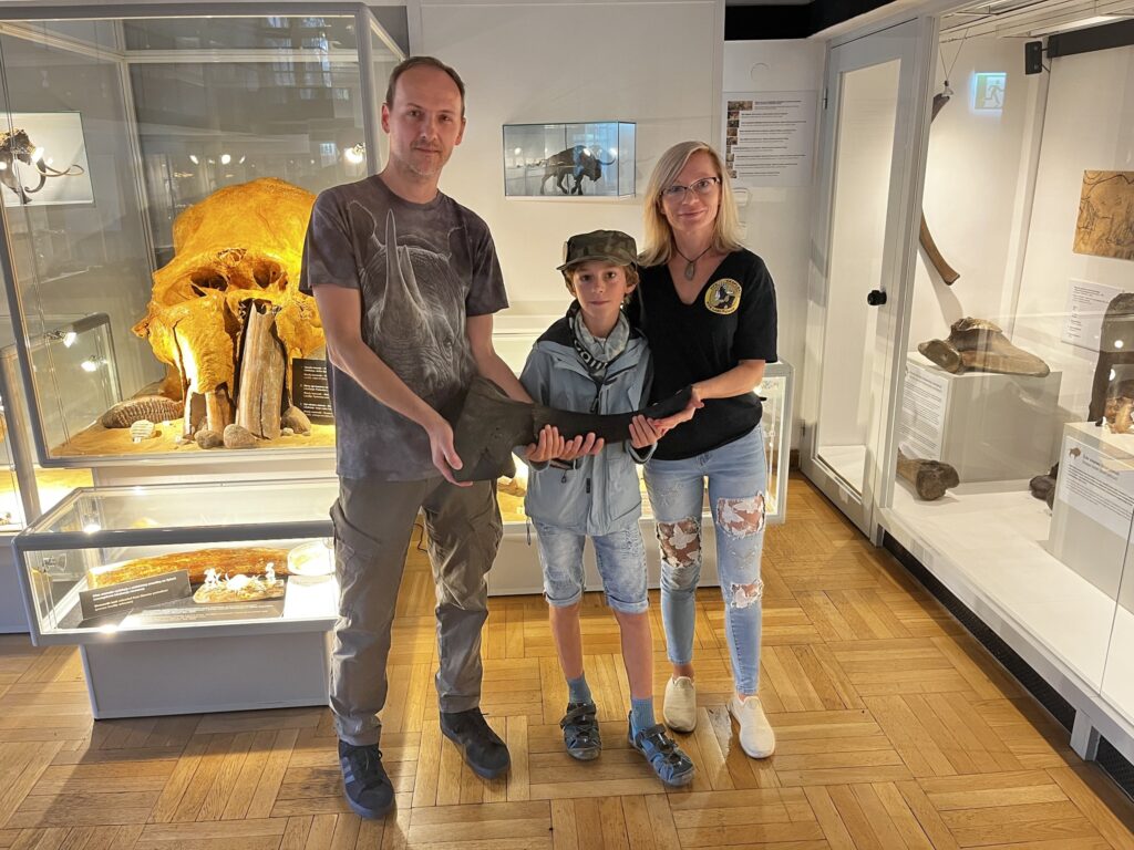 Mężczyzna, chłopiec i kobieta trzymają eksponat w muzeum, w tle widoczne skamieniałości i wystawy.