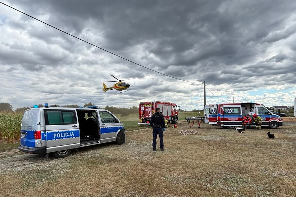 Policjant obserwujący akcję ratunkową w terenie, obok samochodu straży pożarnej i ambulansu, nad którym przelatuje śmigłowiec.