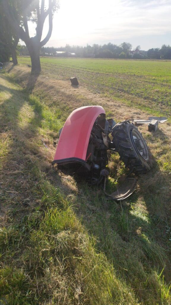 Czerwony fragment ciągnika leży na poboczu wśród trawy, obok widać pole uprawne i drzewa w tle.