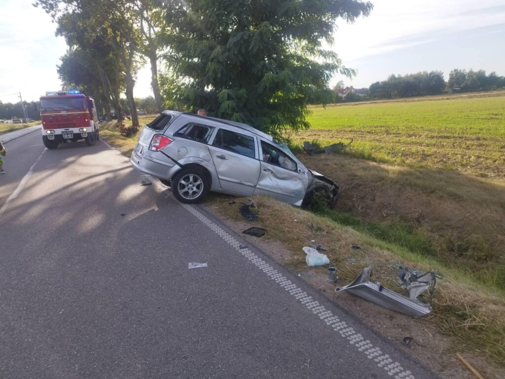 Srebrny samochód osobowy w rowie obok drogi, obok stoi wóz strażacki z włączonymi sygnałami.