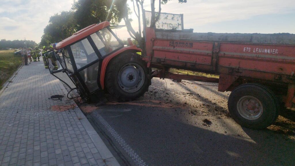 Traktor z przyczepą przechylony na bok na drodze, obok grupka strażaków i ludzi obserwujących sytuację.