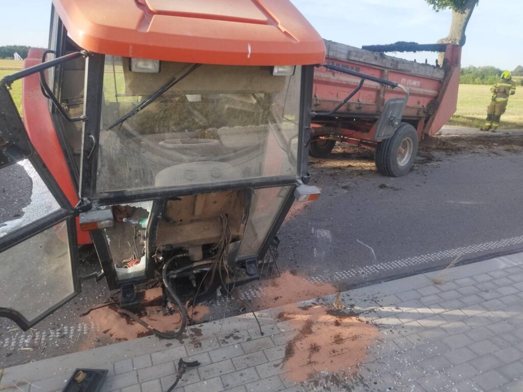 Traktor z uszkodzoną kabiną stoi obok przyczepy, z której wydobywa się błoto, na drodze wiejskiej. W tle widoczny strażak.