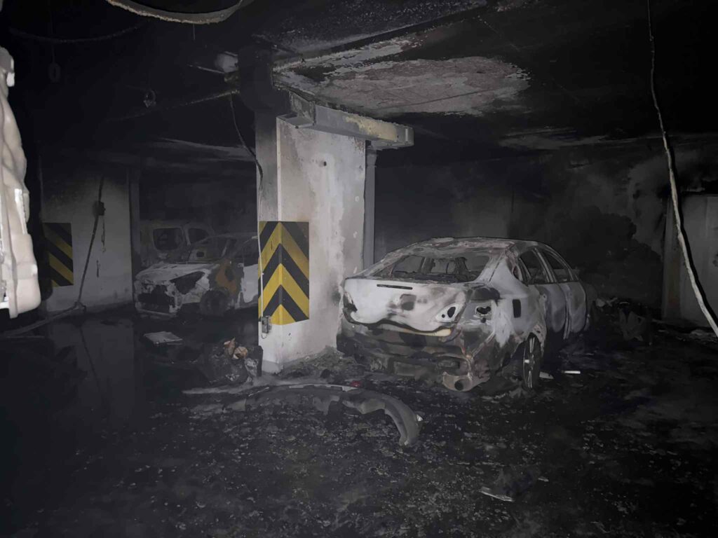 Spalone samochody w ciemnym pomieszczeniu garażowym, z widocznymi oznakami zniszczeń i popiołu.