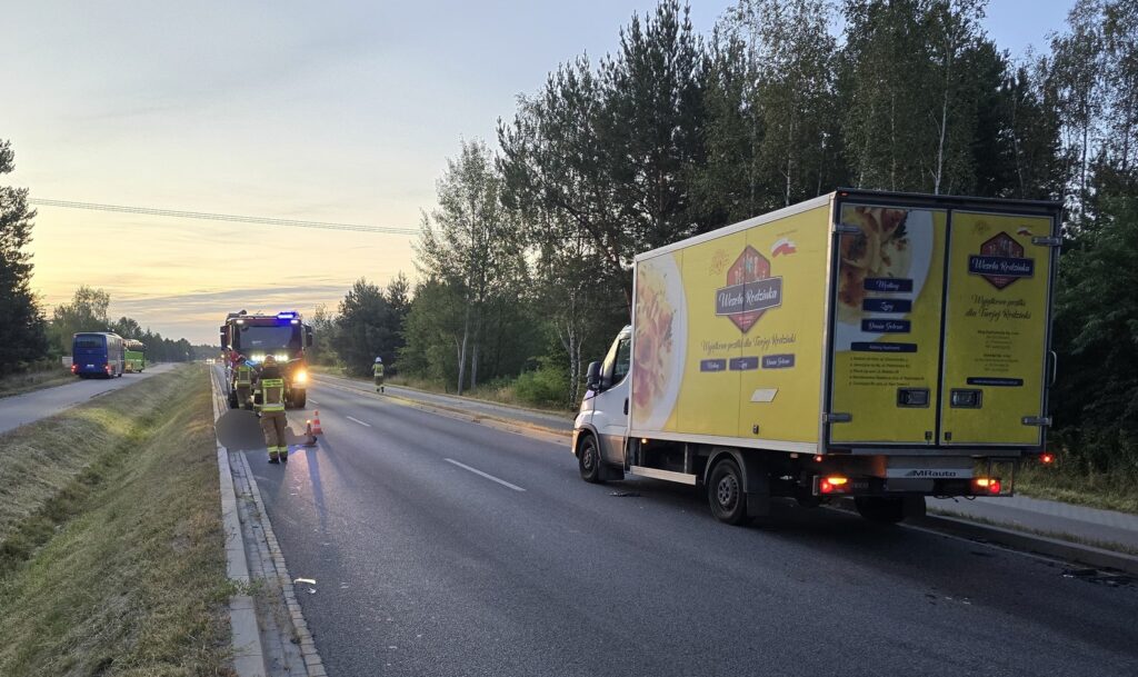 Strażacy pracują przy drodze, obok zaparkowanej ciężarówki z reklamą, w tle wschodzące słońce.