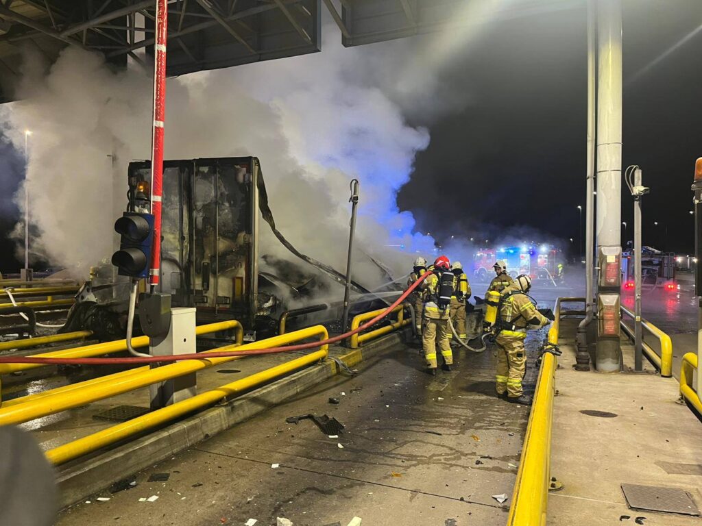 Strażacy gaszą płonący pojazd w otoczeniu dymu na stacji, z włączonymi sygnałami alarmowymi w tle.