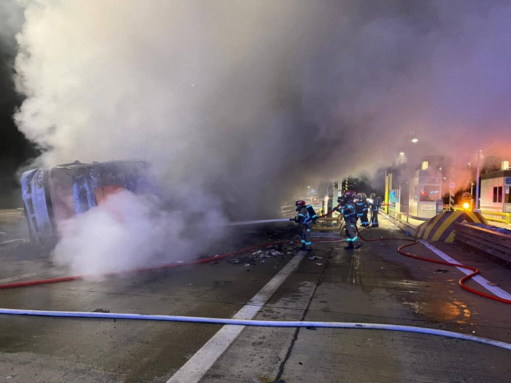 Strażacy gaszą pożar wraku samochodu na drodze, otoczonym gęstym dymem, w nocy przy bramkach opłat.
