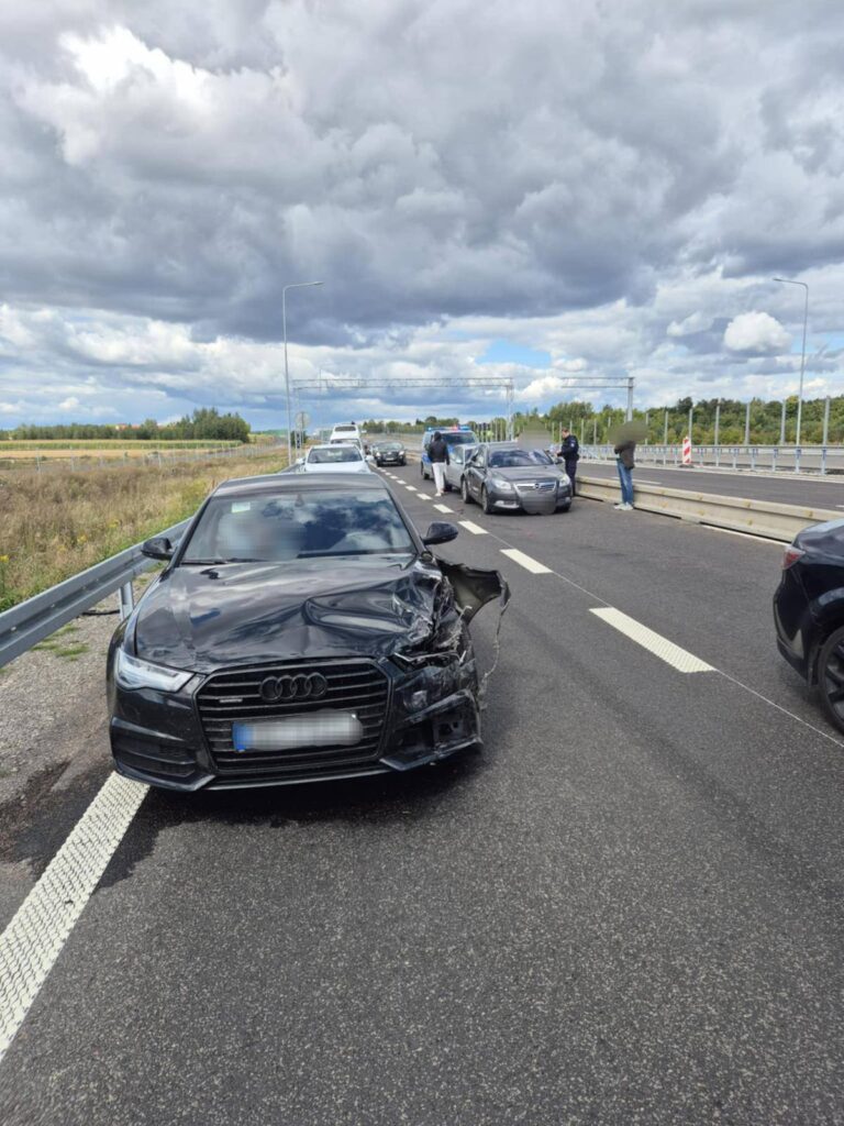 Czarny samochód osobowy z uszkodzoną przednią częścią stoi na drodze, obok grupy ludzi i innych pojazdów.