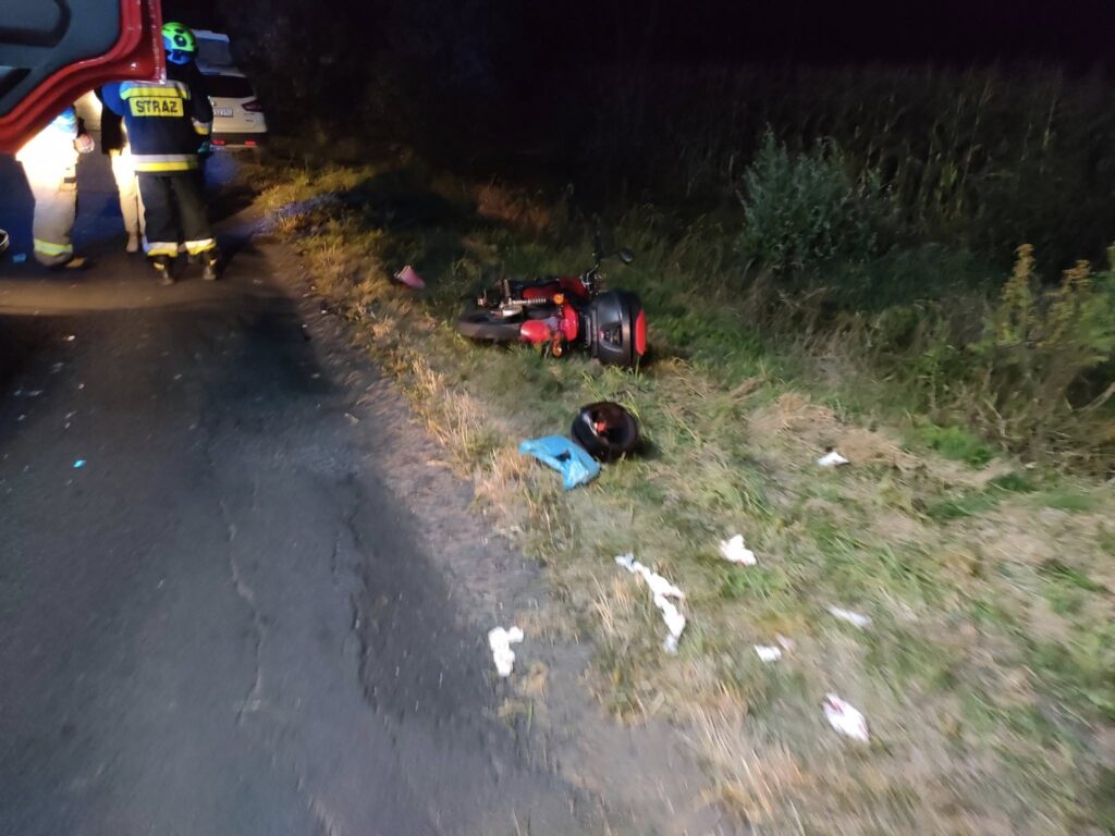 Motocykl leży na poboczu drogi, obok niego kask oraz niebieski przedmiot, a w tle strażacy udzielają pomocy.