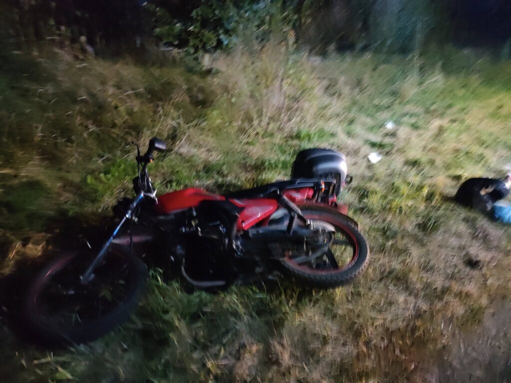 Czerwony motocykl leży na trawie w ciemnym otoczeniu, obok widoczne śmieci i fragmenty otoczenia.
