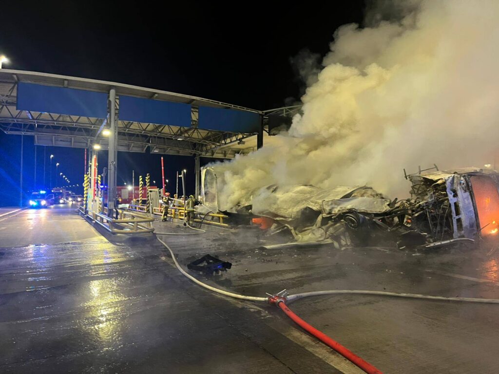 Pojazd w ogniu na stanowisku opłat, otoczony przez strażaków gaszących pożar, w nocy z widoczną mgłą dymu.