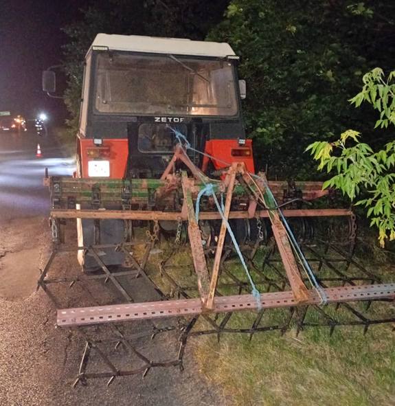 Traktor z zaczepionym narzędziem rolniczym stoi na poboczu drogi w otoczeniu drzew.