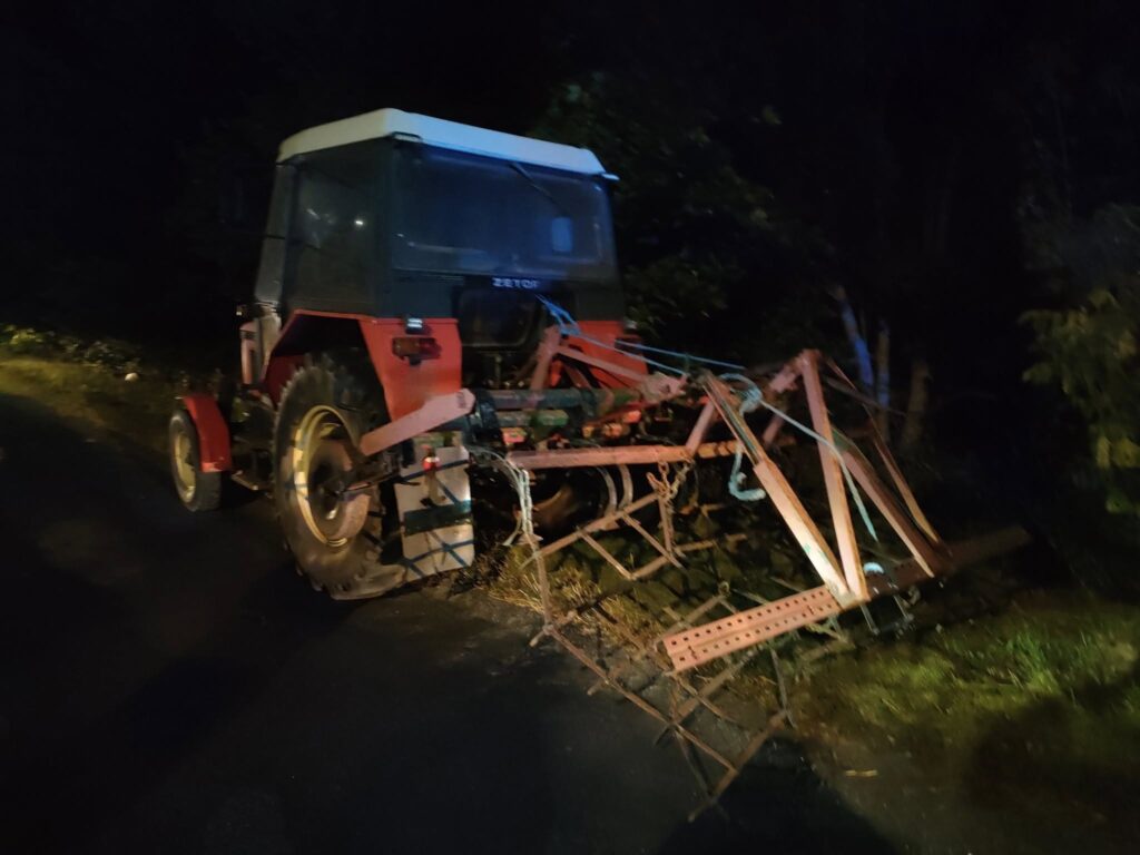 Czerwony ciągnik z przymocowanym sprzętem rolniczym stoi na wąskiej drodze w ciemnym otoczeniu leśnym.