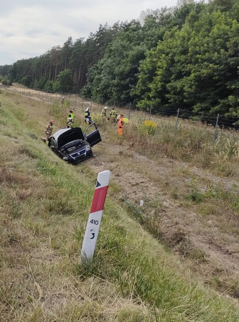 Grupa ratowników w odzieży ochronnej przeszukuje teren przy przewróconym samochodzie na poboczu drogi, otoczonym drzewami.