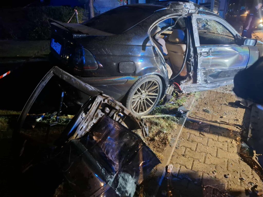 Zniszczony samochód osobowy leży na poboczu drogi, z wyrwaną przednią częścią i otwartymi drzwiami.