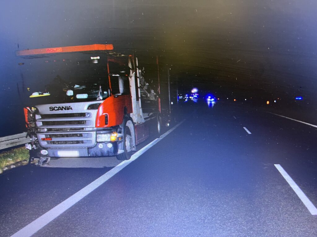 Czerwony ciężarowy pojazd Scania zatrzymany na drodze w nocy, z widocznymi błyskami policyjnych świateł w tle.