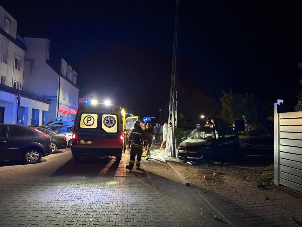 W nocy, na wąskiej ulicy, strażacy oraz zespół ratunkowy interweniują przy uszkodzonym samochodzie, obok karetki.