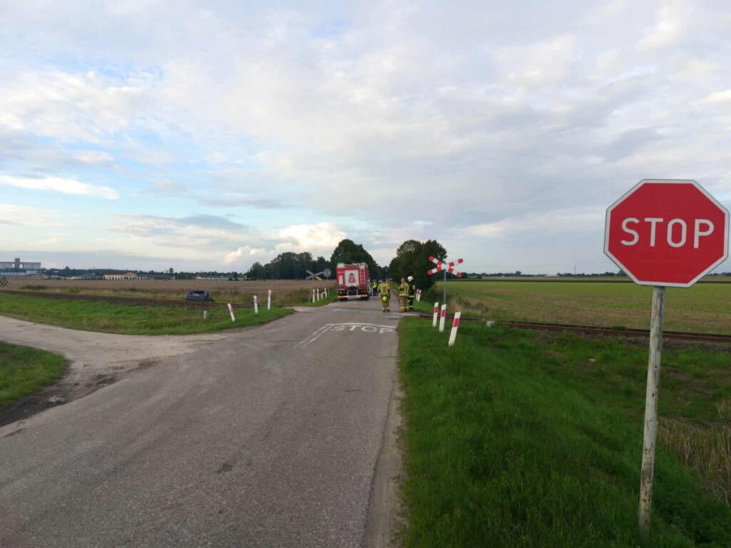 Na skrzyżowaniu drogi z torami kolejowymi stoi znak stop, obok grupy strażaków i wozu strażackiego.