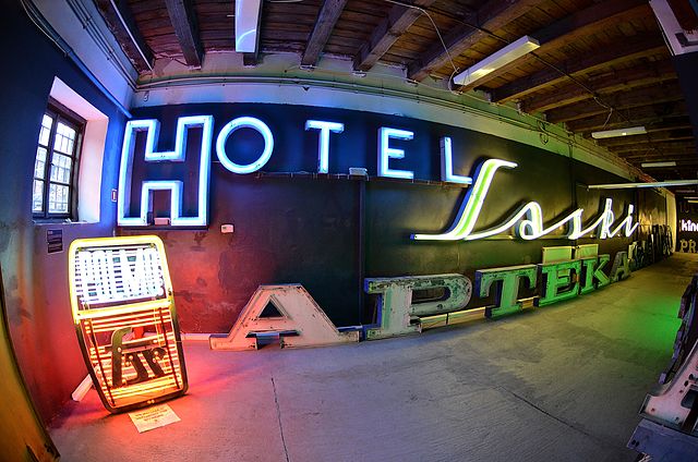 Neonowe znaki "HOTEL" i "APTEKA" na ścianie w industrialnym wnętrzu, oświetlone kolorowymi światłami.