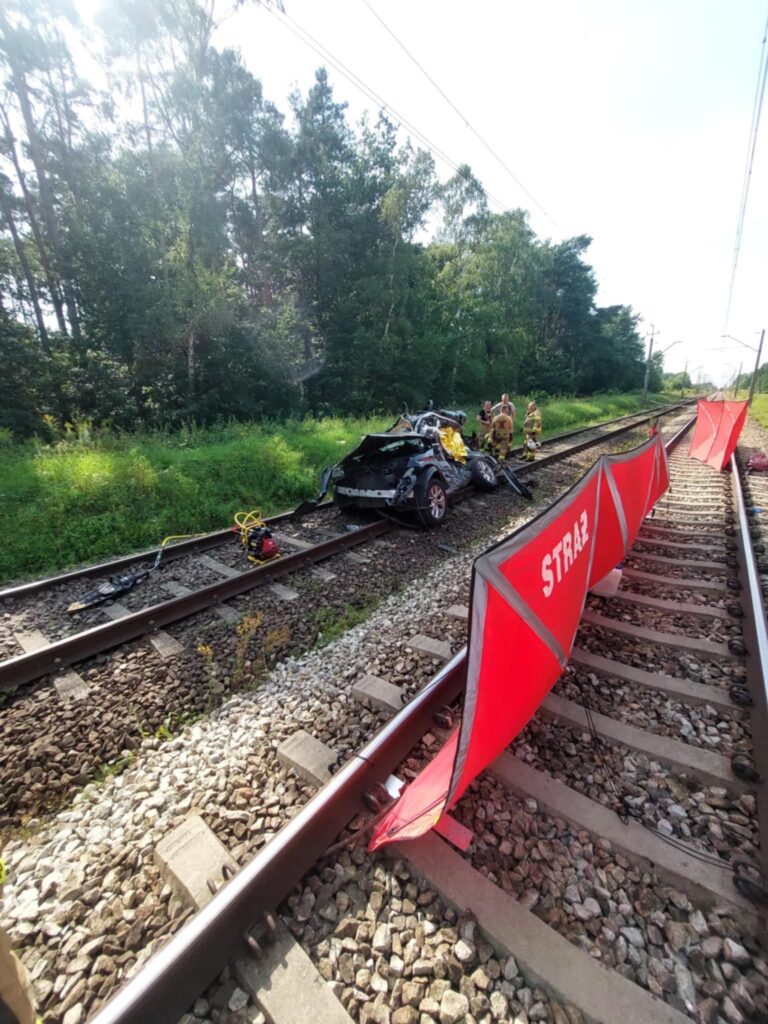 Uszkodzony samochód osobowy leży na torach kolejowych, obok grupy strażaków w mundurach. W tle widoczne drzewa.
