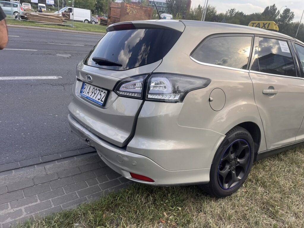 Jasnoszary samochód osobowy zaparkowany na chodniku obok ruchliwej ulicy, z widocznym znakiem taxi na dachu.