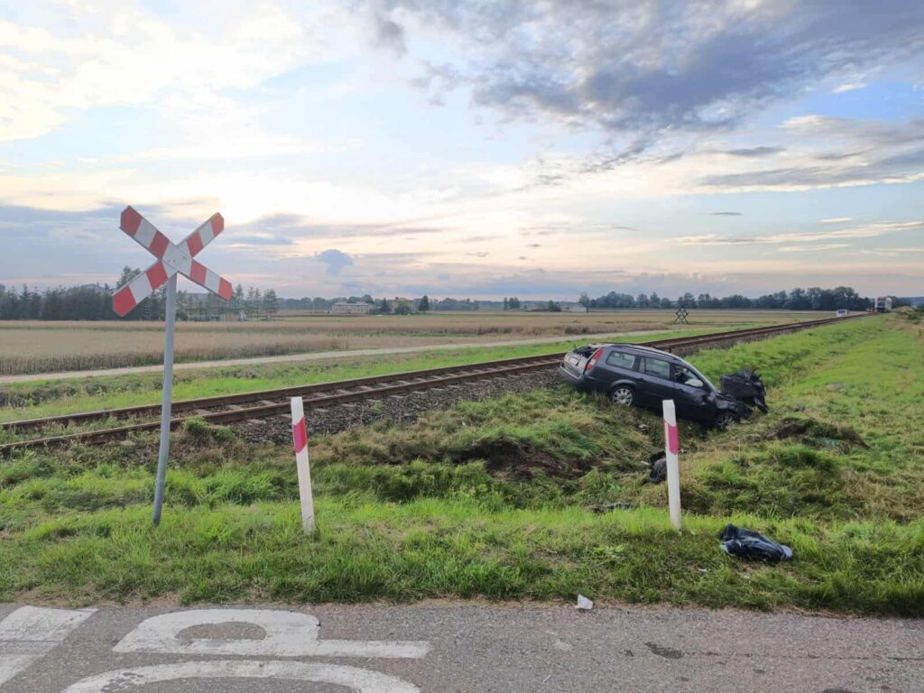 Samochód osobowy w rowie obok torów kolejowych, w otoczeniu pól i chmur na niebie.