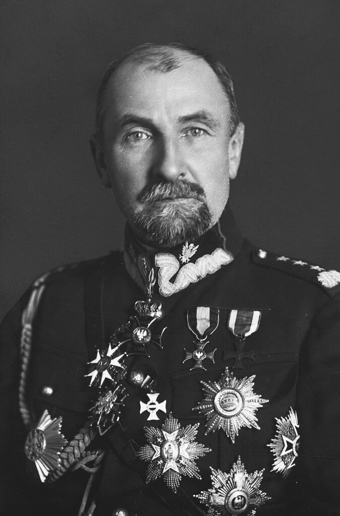 Mężczyzna w mundurze z medalami, z brodą i wąsami, patrzy w obiektyw na ciemnym tle.