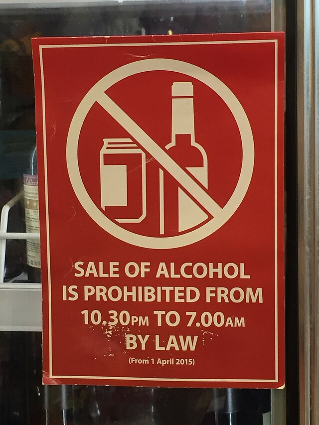 Czerwony plakat informujący o zakazie sprzedaży alkoholu od 22:30 do 7:00, z grafiką przekreślonych butelek i puszek.